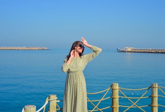 A young beautiful woman in a long dress stands alone on a pier by the sea looking at the sun