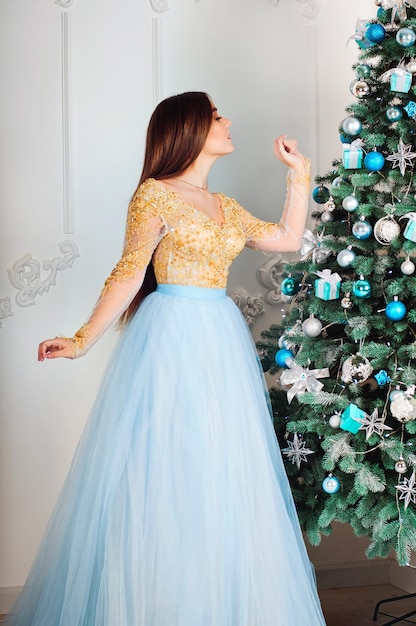 Young and beautiful woman in long party dress sitting near Christmas tree in studio