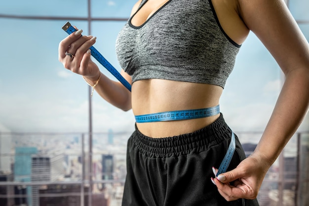 Young beautiful woman measuring perfect slim figure with tape measure in gym with panoramic windows diets Healthy Lifestyle fitness