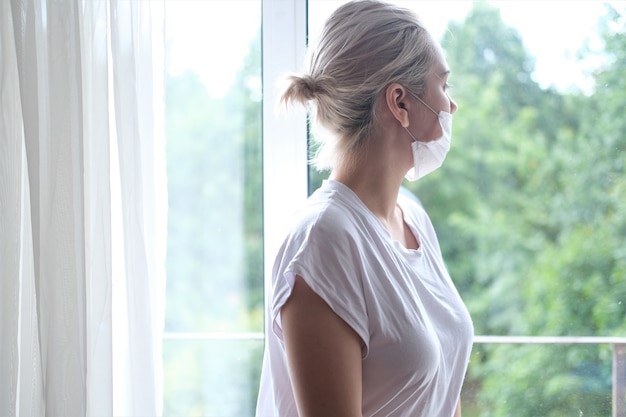 Photo young beautiful woman in a medical mask stands by the window