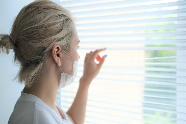 Photo young beautiful woman in a medical mask stands by the window