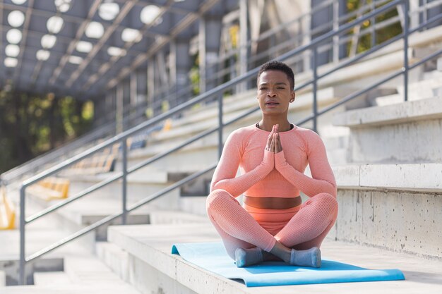 Young beautiful woman meditating in the morning, African American woman dressed in a pink sports suit performs fitness exercises with breathing, near the stadium