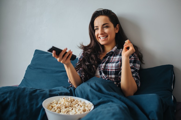 Young beautiful woman in morning bed at home. Positive nice cheerful person watching tv or movie. Enjoying time in bed and eating popcorn.