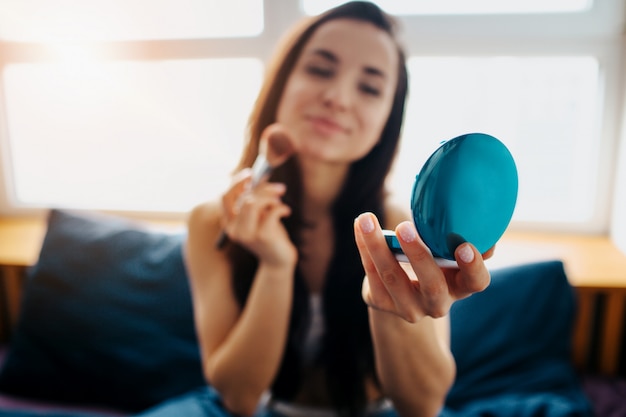 Giovane bella donna nel letto di mattina a casa. brunette attraente sorridente che usando spazzola e polvere con lo specchio. trucco con gioia. tempo di camera da letto.