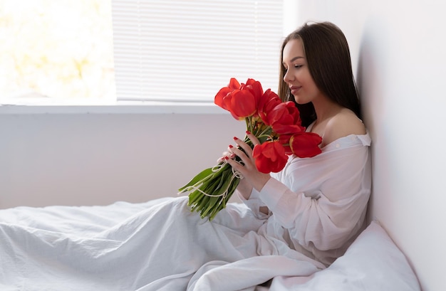 Young beautiful woman in the morning in the bedroom sits with a large bouquet of tulips flowers to a woman for anniversary valentine's day or just like that