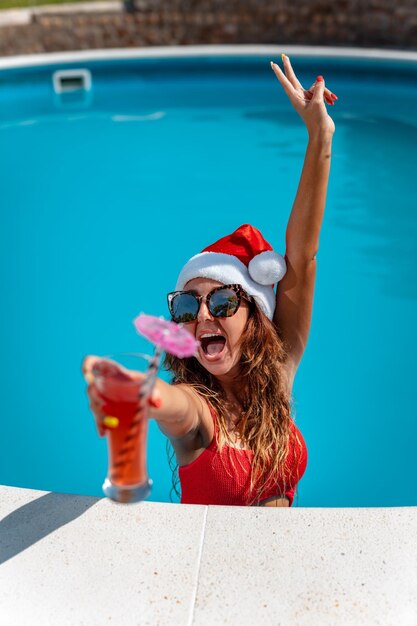 Young beautiful woman near the swimming pool in Santa Claus hat celebrating New Year and Christmas in hot country with glass of cocktail.