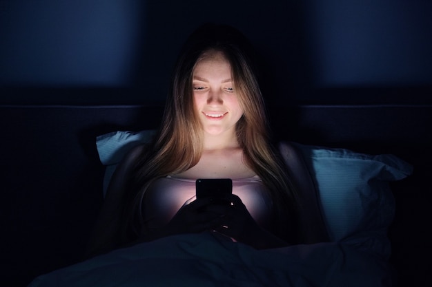 スマートフォンでベッドに座って夜の若い美しい女性