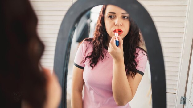 Young beautiful woman in pajamas paints her lips of red lipstick looking in mirror at home Close up of charming female preening before big mirror making makeup