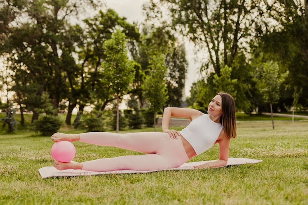 ピンクのスポーツウェアを着た若くて美しい女性が、日の出の公園の芝生の上でピラティスボールを足の間に挟んで横になっているスポーツマットの上に横たわっています。