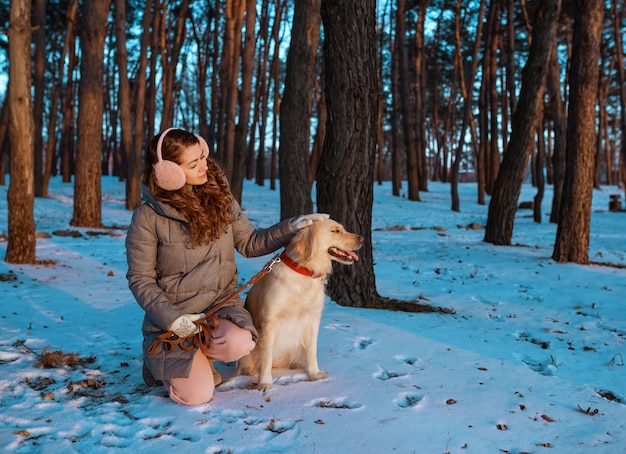 写真 雪の森でゴールデンレトリバー犬をしている若い美しい女性