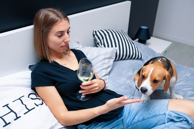 Giovane bella donna che gioca con il cane seduto sul letto in una camera da letto alla moda. donna in una mano che tiene un bicchiere di vino