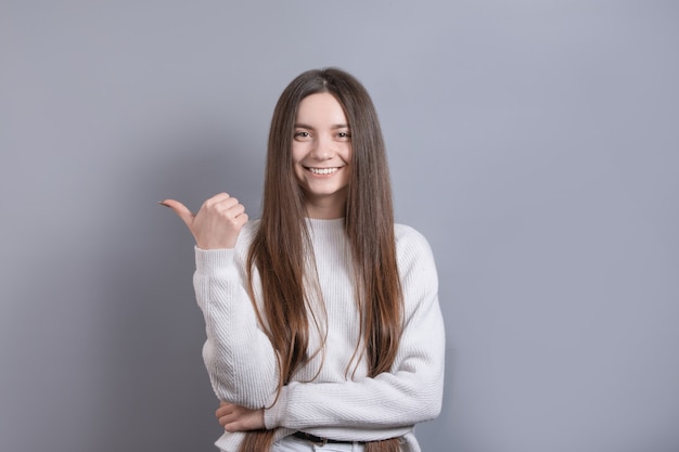 Young beautiful woman portrait with dark hair wearing white sweater smiling with happy face looking and pointing to the side with thumb up.Copy space.