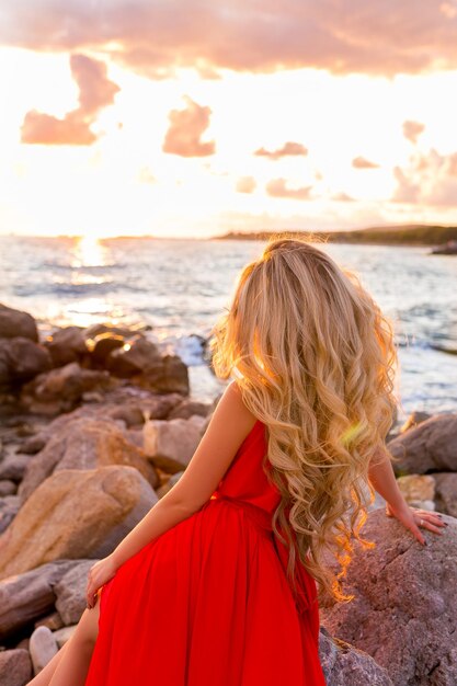 Young beautiful woman in red dress looking to blue sea and fiery sunset