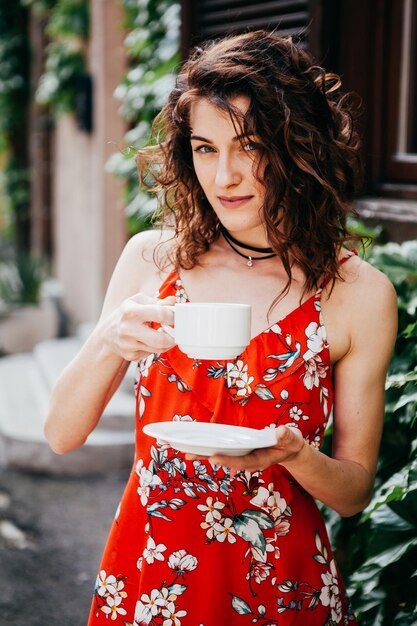 Foto giovane bella donna in un vestito rosso con una tazza sulla strada europea