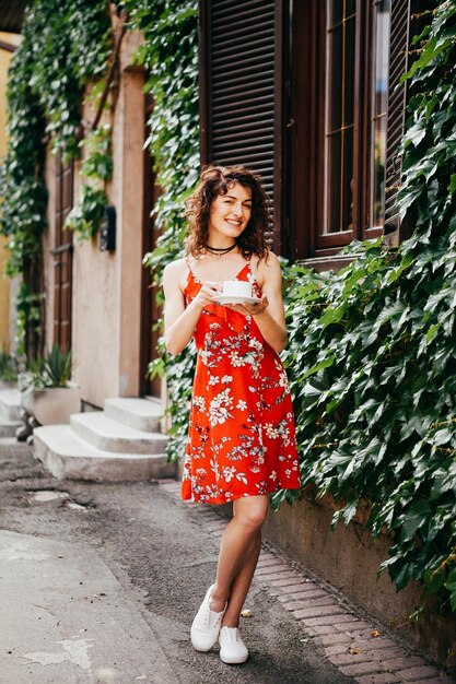 Foto giovane bella donna in un vestito rosso con una tazza sulla strada europea