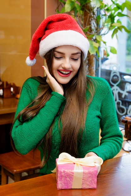 Young beautiful woman in red santa claus hat looking at gift box in cafe