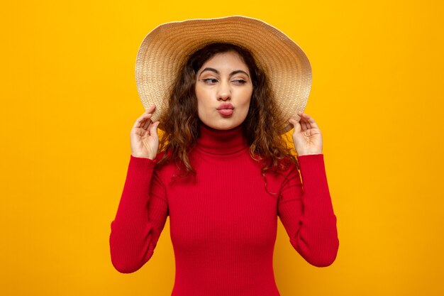 Young beautiful woman in red turtleneck in summer hat looking aside keeping lips like going to kiss standing over orange wall