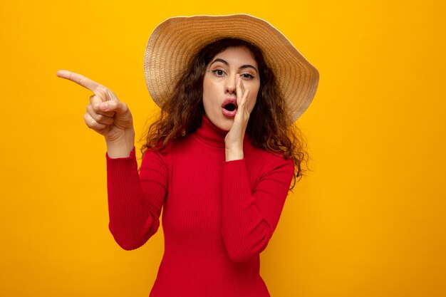 Young beautiful woman in red turtleneck in summer hat looking surprised telling a secret with hand over mouth pointing with finger to the side