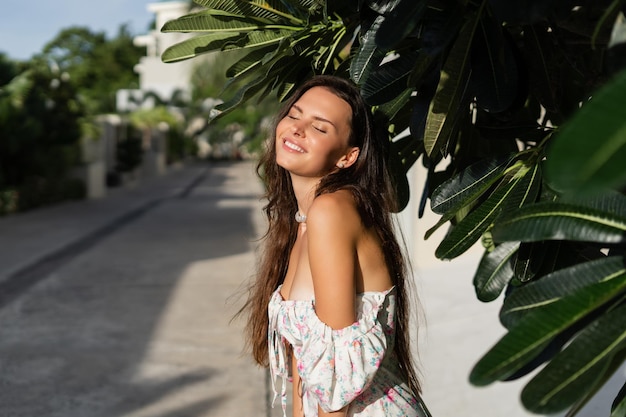 Young beautiful woman in a romantic dress with a floral print and a pearl necklace bracelet against the backdrop of tropical leaves