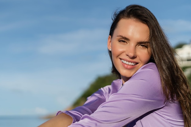 A young beautiful woman in a romantic mood lilac silk clothes on the beach against the backdrop of the sea and stones at sunset laughs smiles with a snowwhite smile