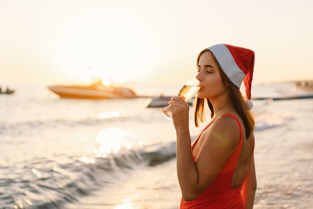 Giovane bella donna con un cappello da babbo natale con un bicchiere di champagne in mano cammina sulla spiaggia