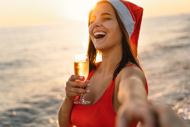 Young beautiful woman in a santa hat with a glass of champagne in her hands walk on the beach