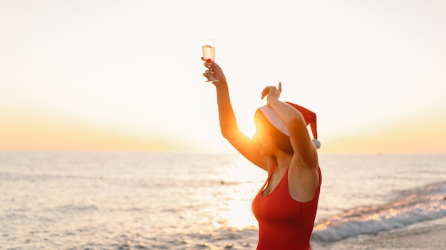 Young beautiful woman in a santa hat with a glass of champagne in her hands walk on the beach