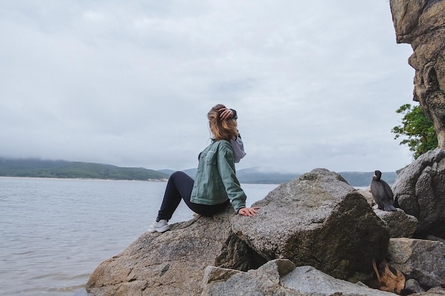 Young beautiful woman sits on the rocks on the seashore and looks into the distance