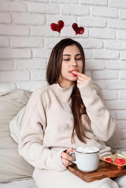 Giovane bella donna seduta nel letto che celebra il giorno di san valentino mangiando dolci e bevendo caffè