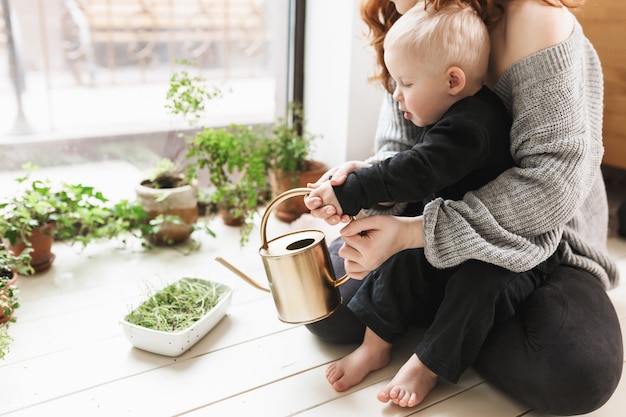 Young beautiful woman sitting on floor with her little son holding watering can in hands with green plants around near big window