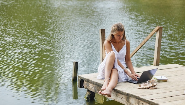 Young beautiful woman sitting at the pier and working on pc