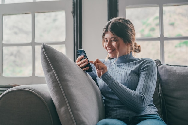 Young beautiful woman sitting on the sofa at home chatting and surfing the net Female person having fun with smartphone online Portrait of girl smiling using cellphone revising social media