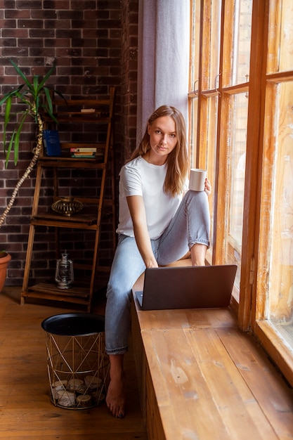 Young beautiful woman sitting at window with a cup of coffee and working with laptop.