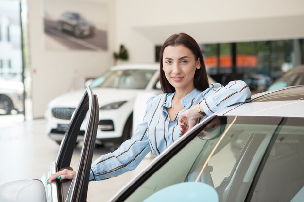 Young beautiful woman smiling to the camera while choosing car to buy at the dealership