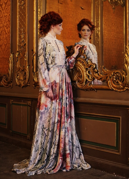 Young beautiful woman standing in the palace room with mirror.