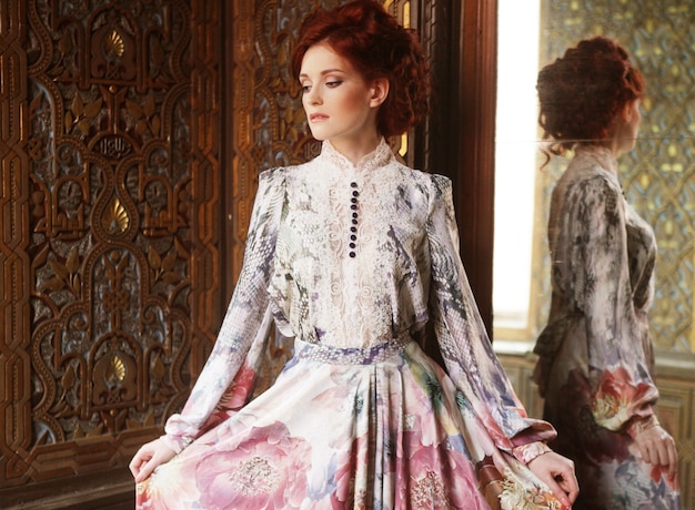 Young beautiful woman standing in the palace room with mirror.