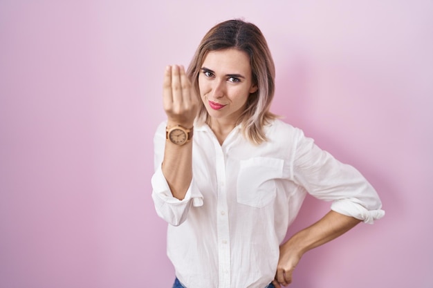 Young beautiful woman standing over pink background doing italian gesture with hand and fingers confident expression