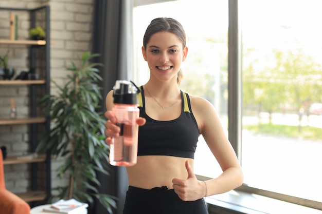 Young beautiful woman standing with dumbbell and drink water after exercising looking camera. Attractive female bodybuilder working out. Fitness and healthy lifestyle concept.