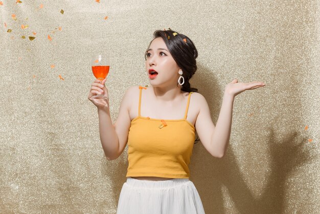 Young beautiful woman standing with glass of champagne in hand and catching confetti over golden glittering background.