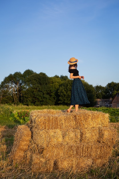 Young beautiful woman in a straw hat stands on a large pile of straw bales