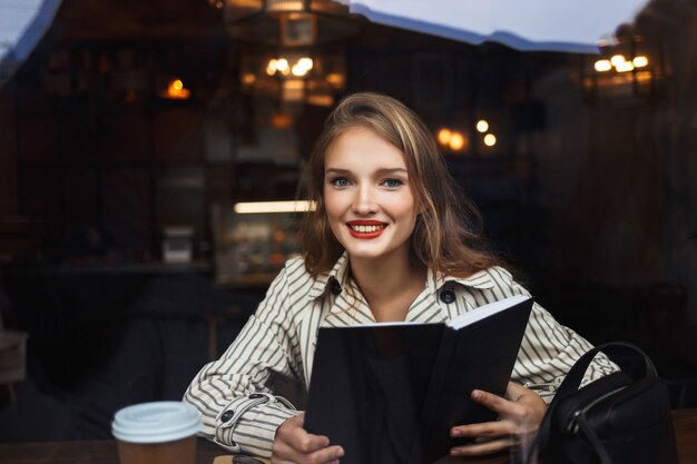 Young beautiful woman in striped trench coat holding book in hands happily 