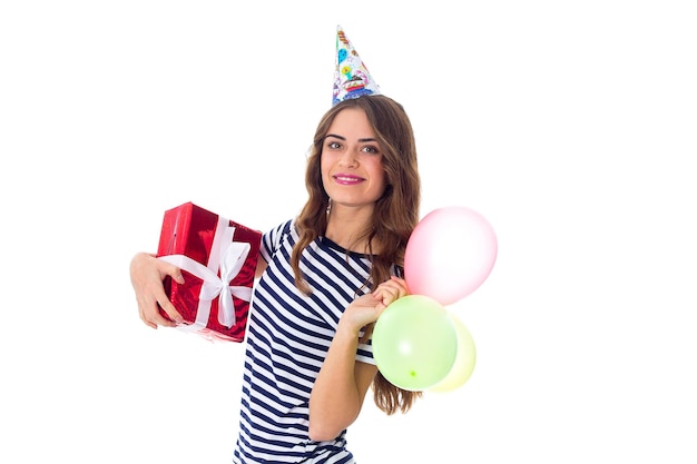 赤いプレゼントと風船を保持しているストリップTシャツとお祝いの帽子の若い美しい女性