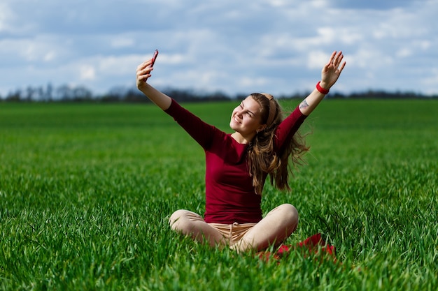 Young beautiful woman student with a phone in her hands sitting on the grass. Girl takes selfies and takes selfie pictures. She smiles and enjoys a warm day. Concept photo on smartphone