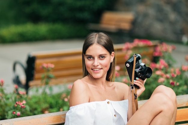 Young beautiful woman in summer clothes holding a retro camera while sitting on bench in the park