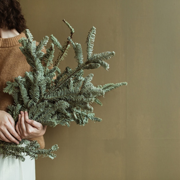 Young beautiful woman in sweater and skirt holding fir branches against olive wall. Minimalist fashion festive Christmas concept.