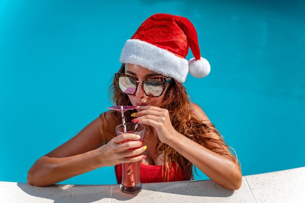Photo young beautiful woman in the swimming pool in santa claus hat celebrating new year and christmas in hot country with glass of cocktail.