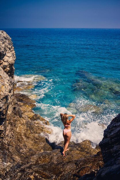 Young beautiful woman in a swimsuit stands on a rocky beach of the Mediterranean Sea The concept of sea recreation Selective focus