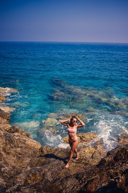 Young beautiful woman in a swimsuit stands on a rocky beach of the Mediterranean Sea The concept of sea recreation Selective focus