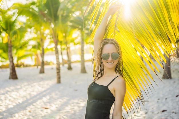 熱帯のビーチで水着を着た若い美しい女性夏休みヤシの木の葉日焼けした肌砂笑顔幸せ幸せな旅行者の女性