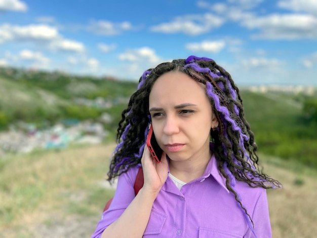 Young beautiful woman talking on mobile phone standing in hilly terrain Cute vivid female with dreadlocks speaking on smartphone standing outdoors in cloudy windy summer weather